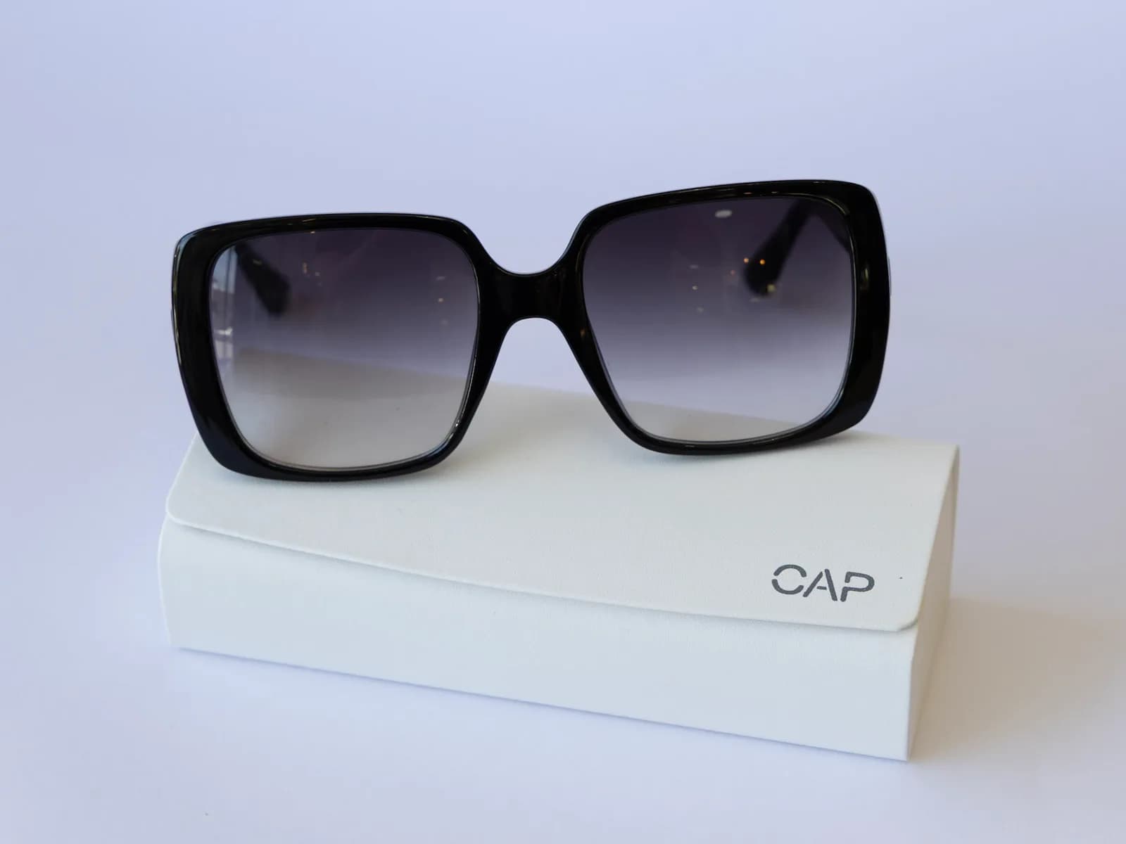 Óculos de sol Noir, quadrado e preto com lentes degradê, apoiado sobre o estojo branco da CAP.